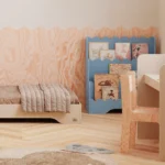 Chambre d’enfant avec un lit blanc et un bureau blanc. Près de la fenêtre se trouve une bibliothèque Lulu bleue à trois étagères, remplie de livres colorés. Mur avec motif en bois et sol clair.