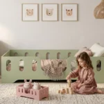 Chambre enfant avec un lit Montessori TILA 2 vert sans barreaux – petite fille jouant avec des blocs à côté d'un lit miniature MINI TILA pour ses peluches