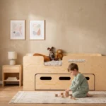 Lit bébé Smartwood PIPI avec tiroir et barrière en bois naturel dans une chambre d'enfant lumineuse - petit garçon jouant avec des cubes en bois à côté du lit.