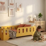 Lit enfant TILA 2 jaune en bois avec barrière dans une chambre claire et apaisante, enfant jouant avec des cubes en bois sur le tapis.