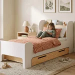 Lit enfant FLOU D avec tiroir, blanc et bois naturel avec étagère à livres, une fillette lit un livre sur le lit.