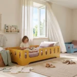 Lit enfant TILA 2 jaune moutarde avec barrières ajourées, conception basse au sol, une fillette lit un livre sur le lit.
