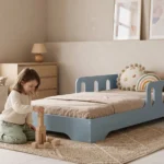 Lit enfant TILA 6 bleu avec barrières ajourées, conception basse au sol, une fillette construit une tour de cubes près du lit.