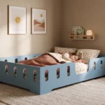 Lit enfant TILA 2 bleu avec barrières, petite fille endormie sous la couverture dans une chambre douce et lumineuse.