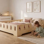 Lit enfant TILA 2 XL en bois naturel avec barrières ajourées, une fillette joue avec des cubes et un nounours près du lit.