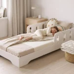 Lit enfant TILA 6 blanc avec barrières, petite fille endormie sur le matelas dans une chambre claire et minimaliste.