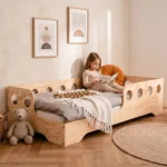 Lit enfant TILA 5 en bois naturel avec barrières à découpes rondes, petite fille lisant un livre appuyée sur un coussin.