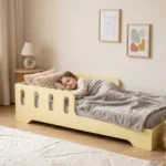 Lit enfant TILA 6 jaune beurre avec barrière, petite fille endormie sous une couverture grise dans une chambre lumineuse.