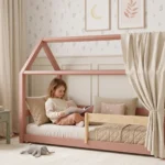 Lit cabane MILO rose avec ciel de lit en tissu et barrière, petite fille assise qui lit un livre dans sa chambre fleurie.