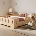Lit enfant TILA 2 coloris naturel avec barrières ajourées, petite fille assise sur le tapis qui lit un livre à côté du lit.