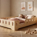 Lit enfant TILA 2 coloris naturel avec barrières ajourées, petite fille endormie avec sa peluche sous une couette fleurie.