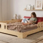 Lit enfant TILA 1 coloris naturel avec barrières ajourées sur trois côtés, petite fille assise qui lit un livre adossée aux coussins.