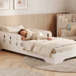 Lit enfant TILA 6 blanc avec barrière ajourée, petite fille endormie sous une couette beige dans une chambre lumineuse.